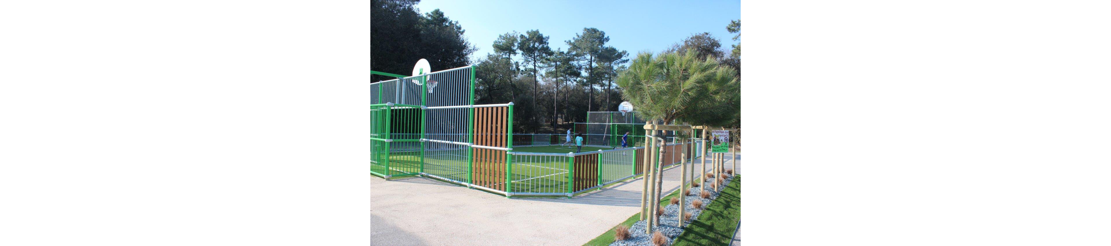 An outdoor multi-sport court with green and white fencing is set within a landscaped park. Children play on the artificial turf field, with basketball hoops mounted above the high metal barriers. Tall trees and a clear sky create a bright, natural backdrop around the sports area. A paved path and neatly planted shrubs line the edge of the court, adding to the organized recreational setting.
