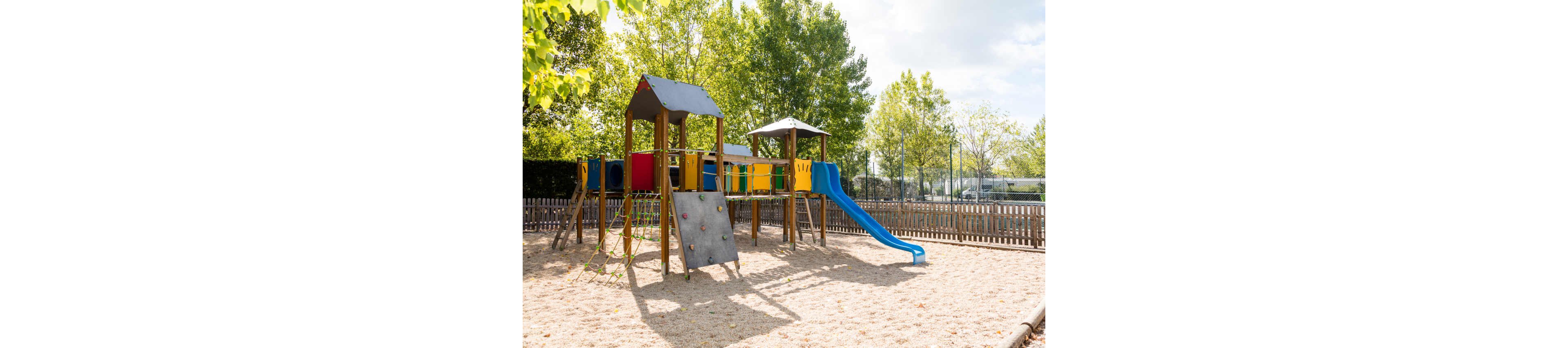 A colorful wooden playground structure stands in a sandy play area. The equipment includes towers, a blue slide, a climbing wall, and rope elements. A wooden fence surrounds the playground, with trees and a sports court visible in the background. Bright daylight and green foliage create a cheerful, family-friendly atmosphere.