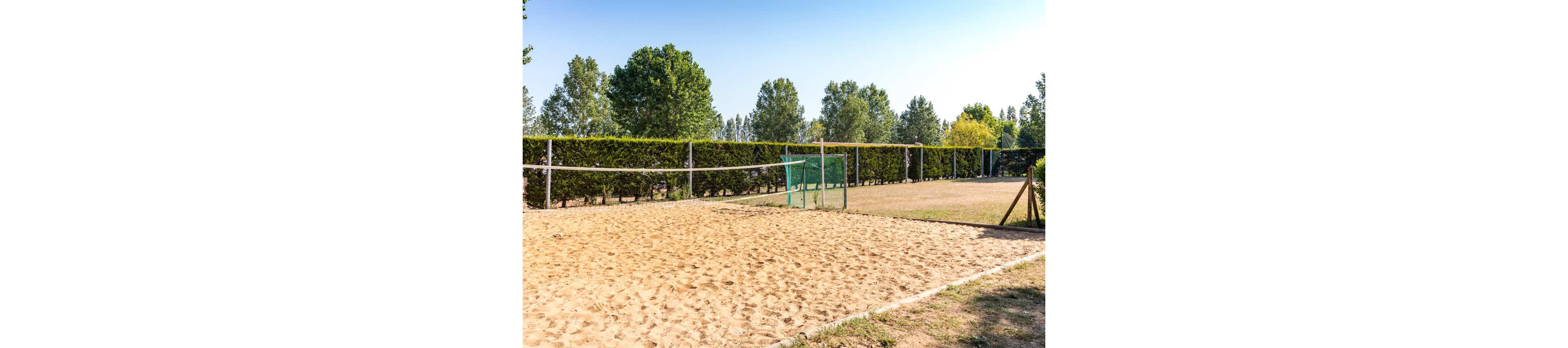A sandy volleyball court is set up outdoors with a net stretching across the middle. The court is surrounded by green hedges and tall trees under a clear blue sky. A small green fence or barrier stands near the center of the court area. The scene suggests a recreational sports space in a quiet, natural setting. Bright sunlight and shadows emphasize the warm, dry ground and relaxed atmosphere.