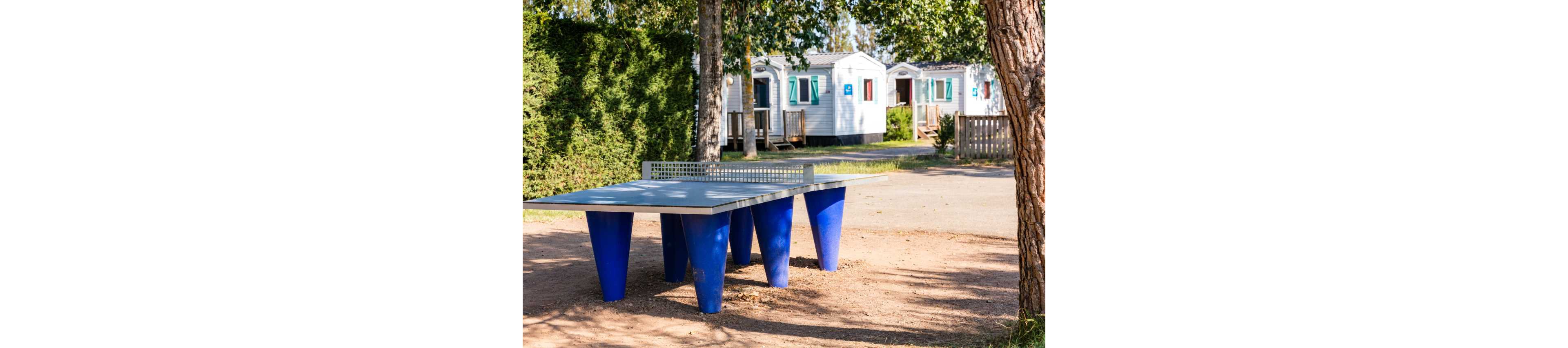 A modern outdoor ping pong table with bright blue legs stands in a sunny campsite. Behind it, several white mobile homes with colorful shutters line a quiet lane. Trees and hedges provide shade and greenery around the recreational area. The scene suggests a relaxed vacation setting with space for outdoor games and leisure.