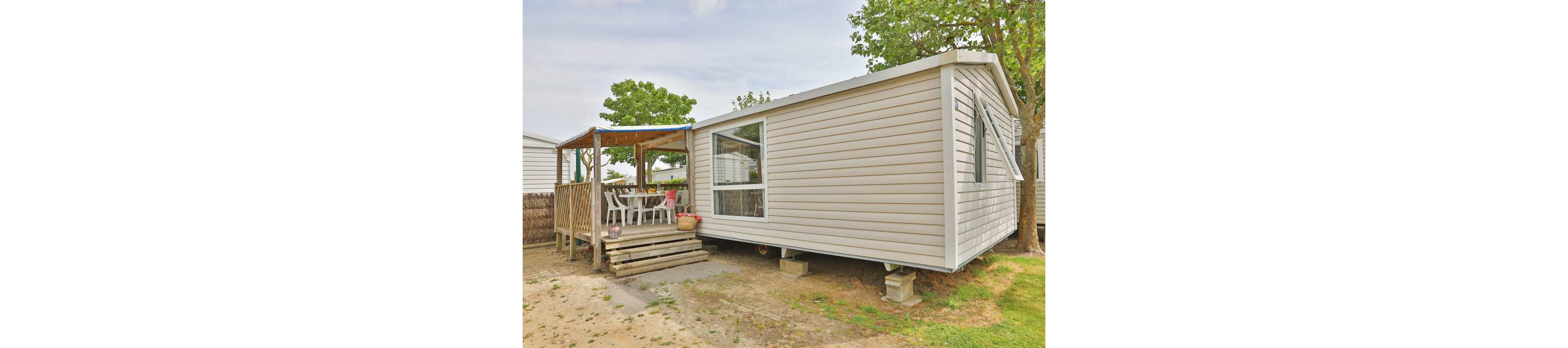 A light-colored mobile home stands elevated on concrete blocks in an outdoor setting. A covered wooden deck extends from the side, furnished with a dining table and several chairs for outdoor meals. Large windows on the home allow natural light inside, while trees and neighboring structures suggest a campsite or holiday park. The ground around the home is a mix of grass and bare soil, giving a relaxed, rustic atmosphere.