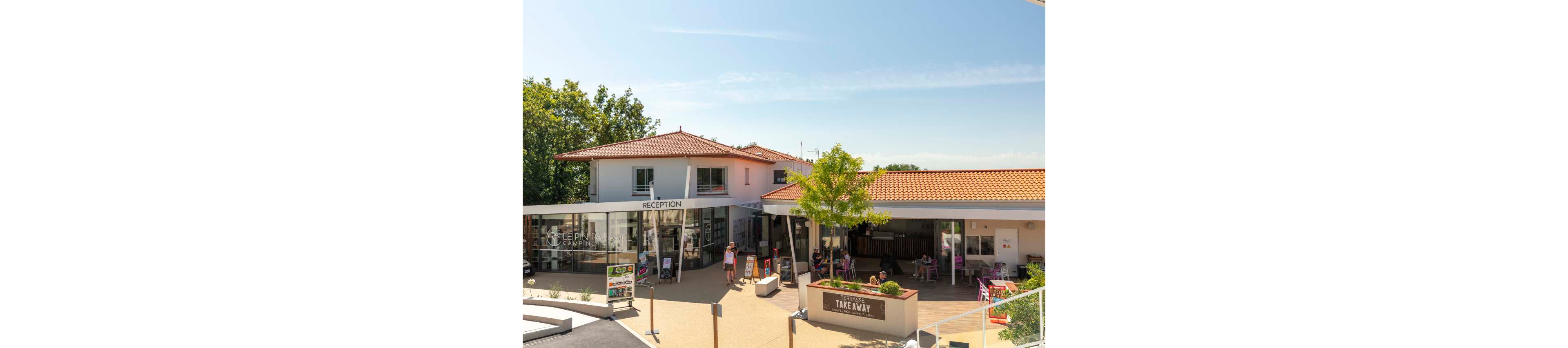 A modern campsite reception building with red-tiled roofs is shown under a clear blue sky. Guests walk and relax around the entrance area, where large glass windows reveal the interior. A separate terrace area labeled takeaway features outdoor seating shaded by a tree. The scene conveys a relaxed holiday atmosphere in a well-maintained vacation facility.