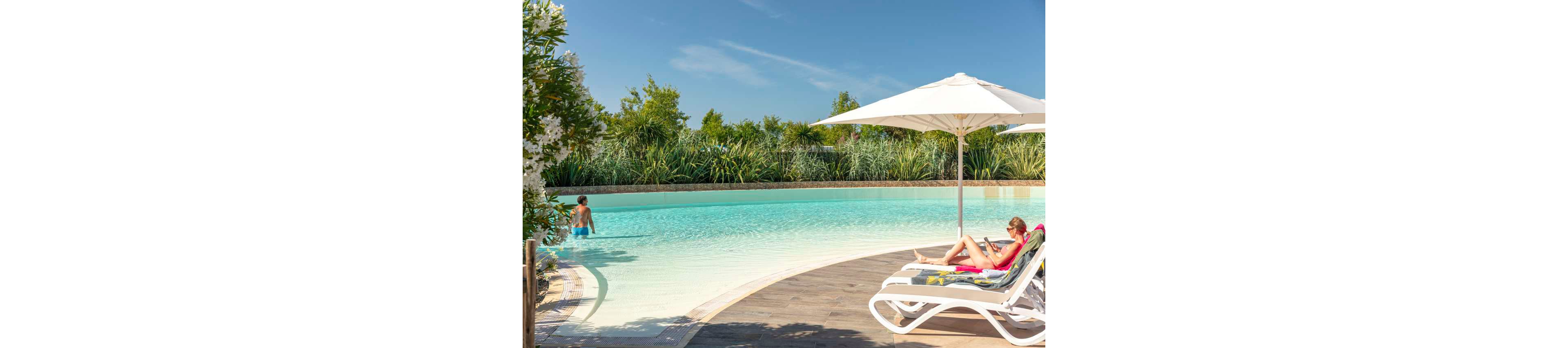 A woman lounges on a white sunbed under a large umbrella beside a curved outdoor swimming pool. Another person stands in the shallow water near the pool’s edge. Lush green plants and trees surround the pool, creating a secluded resort atmosphere. The bright sunlight, clear blue sky, and calm water convey a peaceful summer vacation mood.