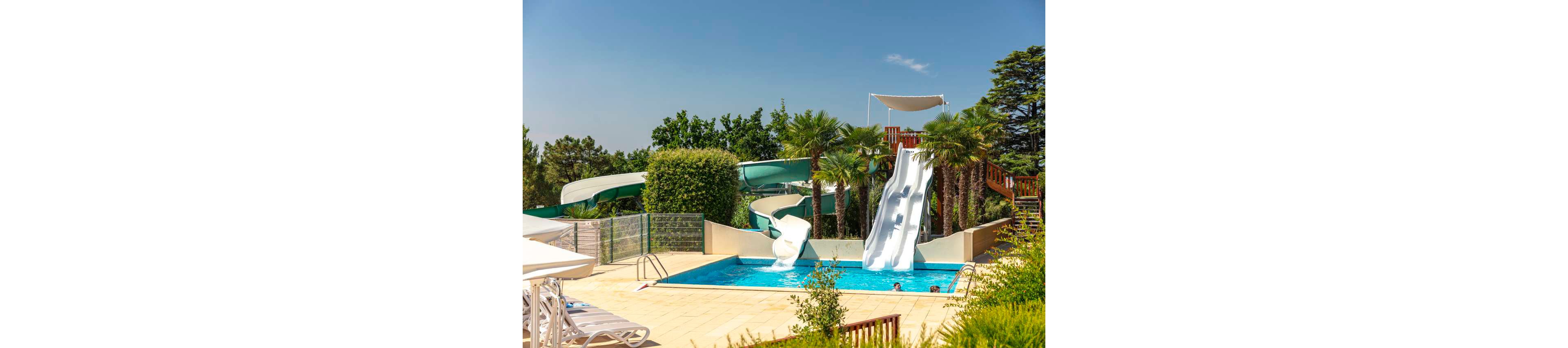 An outdoor swimming pool features multiple large water slides ending in the clear blue water. The scene is set in a sunny resort area surrounded by palm trees, lush greenery, and a wooden staircase leading to the slide platforms. A few people are visible in the pool enjoying the water. Lounge chairs and umbrellas line the tiled pool deck, suggesting a relaxed vacation atmosphere.