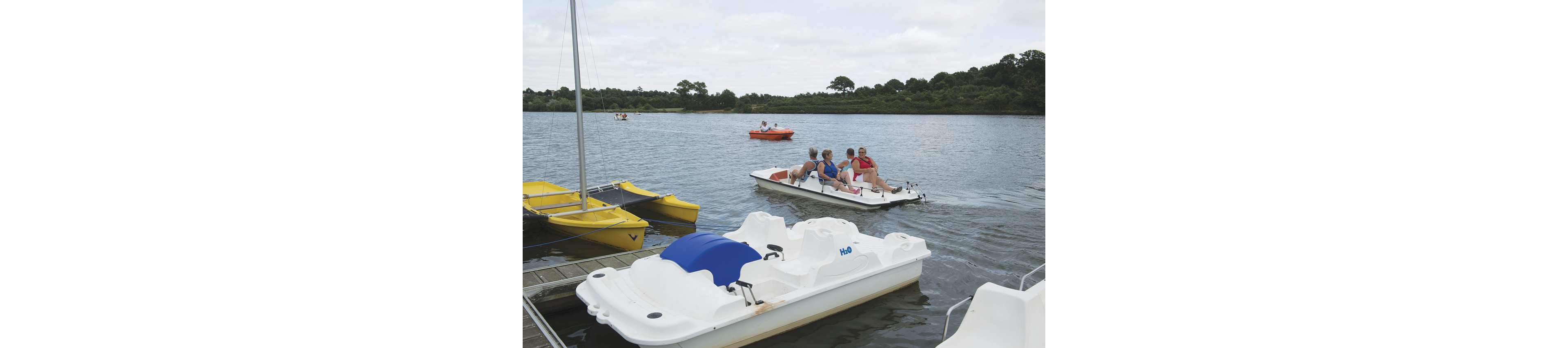 A group of adults ride a white pedal boat together on a calm lake. Several other small boats, including yellow and orange ones, float in the background near a tree-lined shore. An empty white pedal boat with a blue cover is docked in the foreground. The scene suggests a relaxed recreational outing on the water under a cloudy sky.