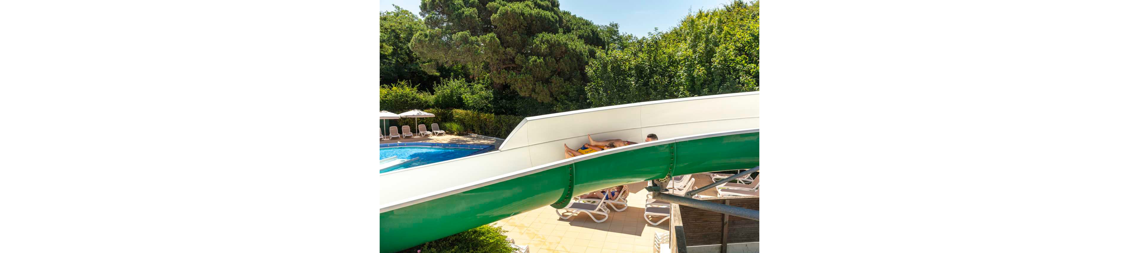 A group of people ride together on a large green waterslide at an outdoor pool. The slide curves above a sun terrace with empty lounge chairs and umbrellas. A clear blue swimming pool is visible to the left, surrounded by lush green trees and bushes. The bright sunlight and shadows suggest a warm summer day at a leisure resort or water park.