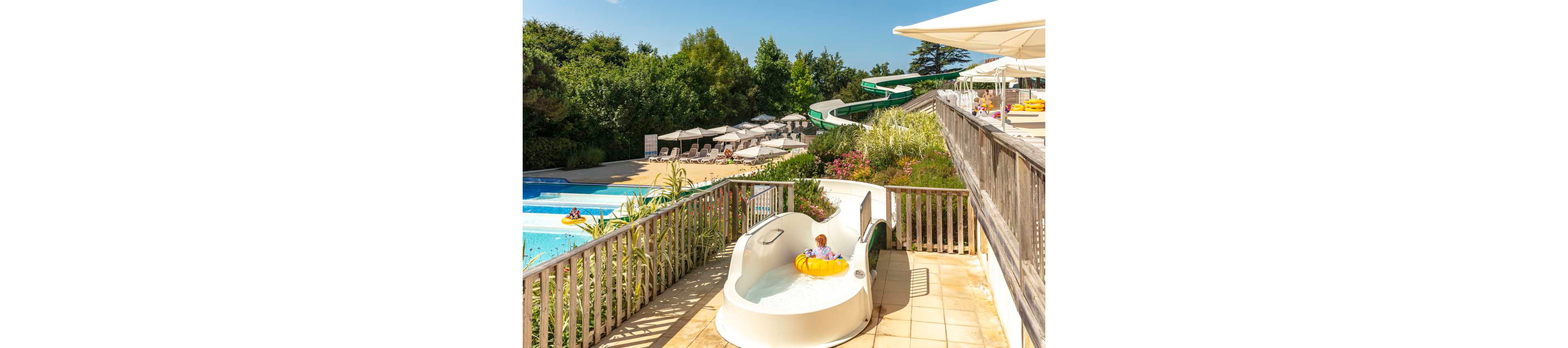 A young child rides a yellow inflatable ring down a white waterslide at an outdoor resort pool. The slide curves toward a bright blue swimming pool where other guests relax. Rows of sun loungers and umbrellas line the poolside terrace under clear, sunny skies. Lush green trees and landscaped plants surround the water park area, creating a vibrant holiday atmosphere.
