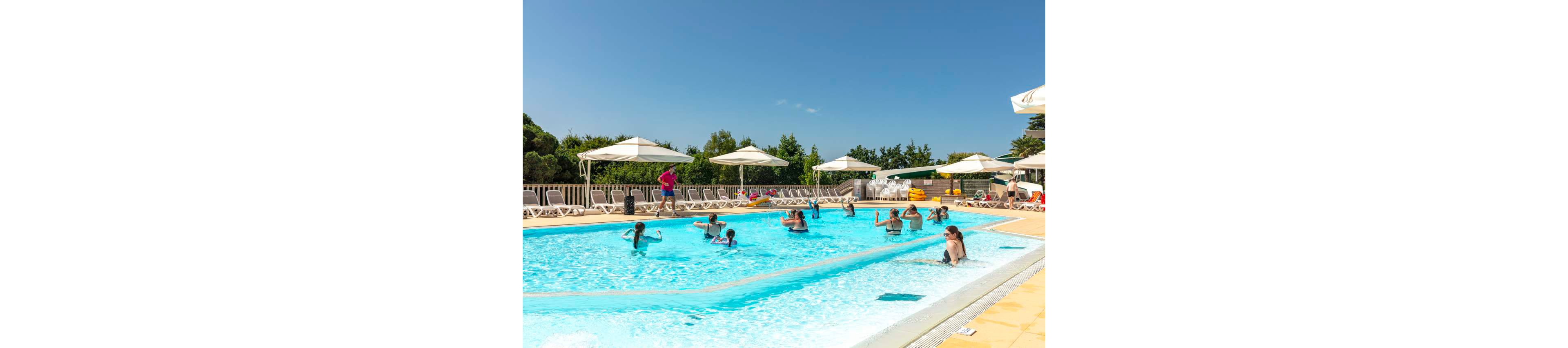 A group of people participate in a water aerobics class in a bright outdoor swimming pool. The instructor stands at the poolside while adults and children follow movements in the water. White sun loungers and large umbrellas line the deck, suggesting a resort or holiday setting. The sky is clear and blue, creating a relaxed and summery atmosphere.