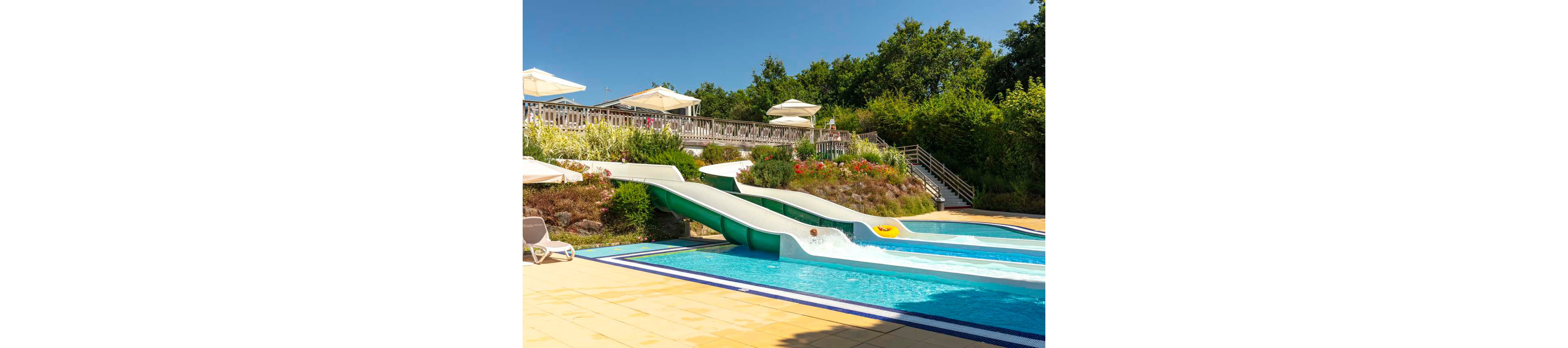 An outdoor swimming pool features two parallel green water slides ending in clear blue water. A person on a yellow inflatable ring splashes into the pool while another slide lane is empty. Sun loungers and large white umbrellas line the deck above, surrounded by landscaped greenery and colorful flowers. A wooden fence and stairway lead up to the terrace area, with dense trees in the background under a bright blue sky.