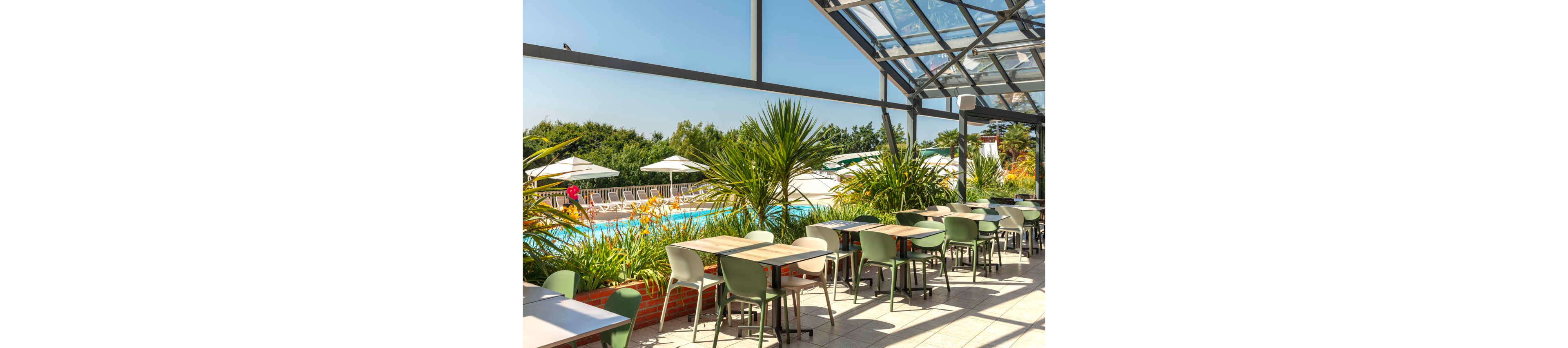 An outdoor terrace café overlooks a bright blue swimming pool on a sunny day. Rows of modern tables and chairs sit beneath a partial glass roof, creating a relaxed dining area. Lush green plants in planters separate the seating from the pool deck. White sun umbrellas and lounge chairs are visible in the background, framed by trees and clear blue sky.