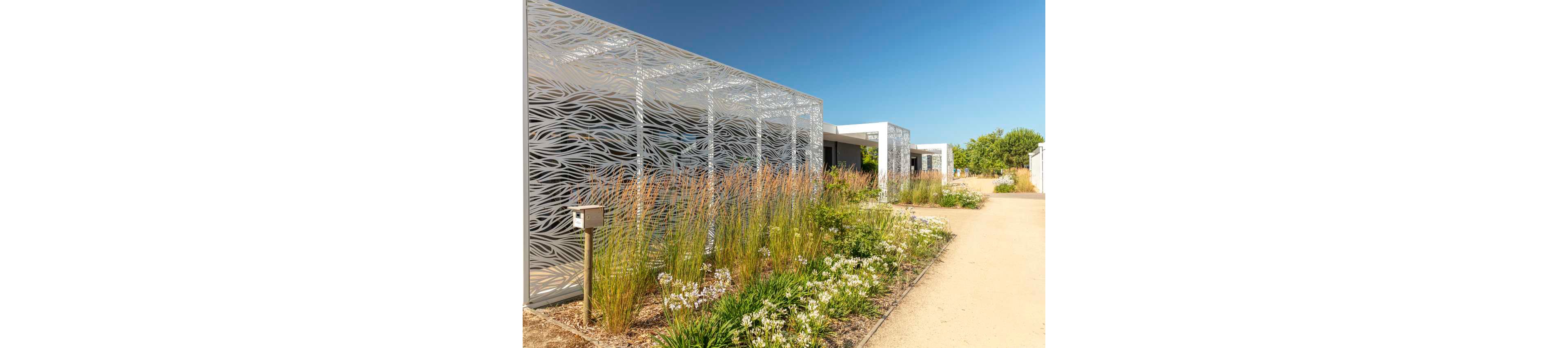 A contemporary outdoor walkway features tall decorative metal screens with an abstract wavy cutout pattern. The path runs alongside a landscaped garden filled with ornamental grasses and flowering plants. A modern building with clean white lines is visible in the background under a clear blue sky. The scene conveys a bright, airy atmosphere in a well-designed architectural landscape.