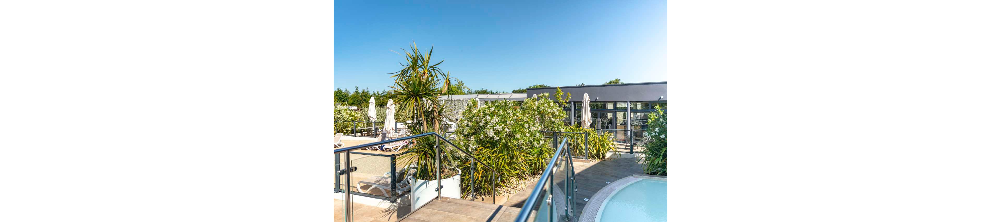 A contemporary outdoor pool area features a curved swimming pool beside a raised sun terrace. Glass and metal railings frame the steps leading up to loungers and umbrellas. Lush green plants and small palm trees add a tropical feel to the modern building in the background. The bright blue sky and strong sunlight create a warm, inviting resort atmosphere.