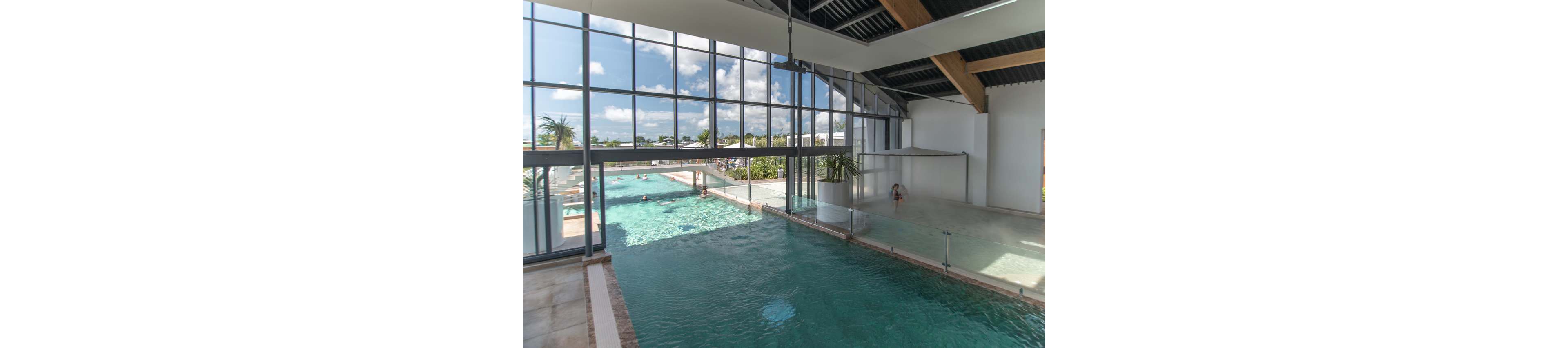 A large indoor swimming pool opens onto an outdoor pool through expansive glass walls. Sunlight streams in through the windows, reflecting off the clear blue water and polished floor. A few people relax and swim in the outdoor section, while the indoor area appears calm and spacious. The modern structure features high ceilings, exposed beams, and tropical plants visible outside.