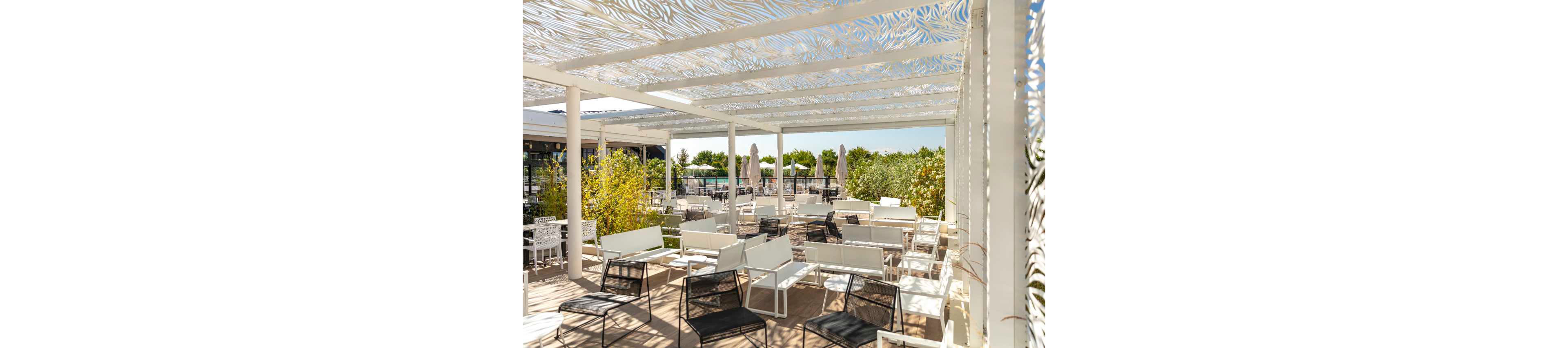 An open-air terrace features modern white and black chairs and tables arranged on a wooden deck. A decorative white pergola with intricate cutout patterns casts dappled light across the seating area. Green shrubs and plants border the space, with large umbrellas visible in the background near a pool or garden area. The bright daylight and clean design create a relaxed, contemporary outdoor lounge atmosphere.