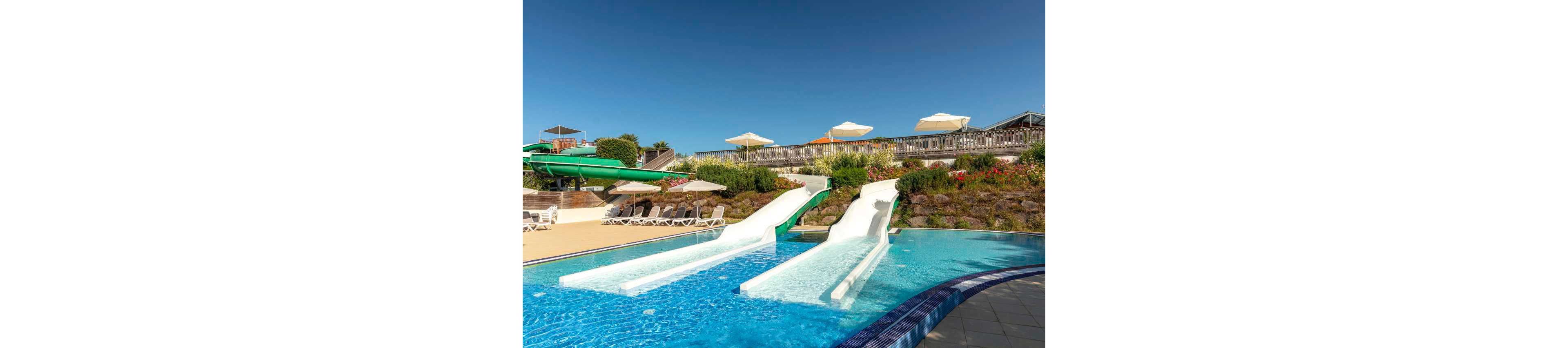 An outdoor swimming pool features multiple water slides ending in clear blue water. Two white straight slides and a green tube slide descend from a landscaped terrace. Sun loungers and large white umbrellas line the poolside area under a bright blue sky. Colorful flowers and greenery add a resort-like atmosphere to the scene.