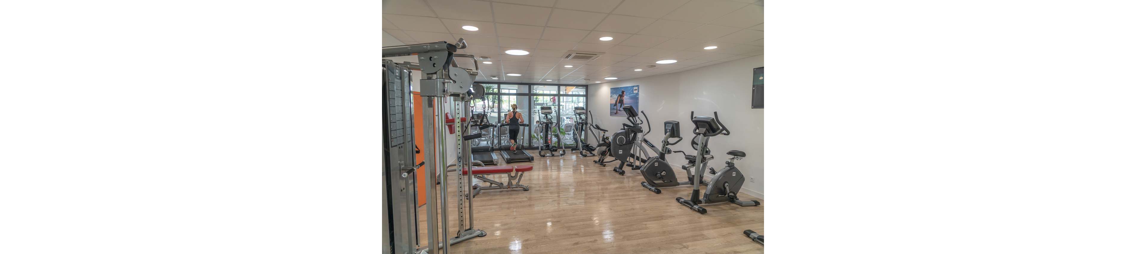 A bright, modern gym features various exercise machines arranged across a polished wooden floor. A person runs on a treadmill near large windows that let in natural light. Stationary bikes, weight machines, and strength-training equipment are visible throughout the room. The clean, spacious interior creates an inviting environment for indoor workouts and fitness training.