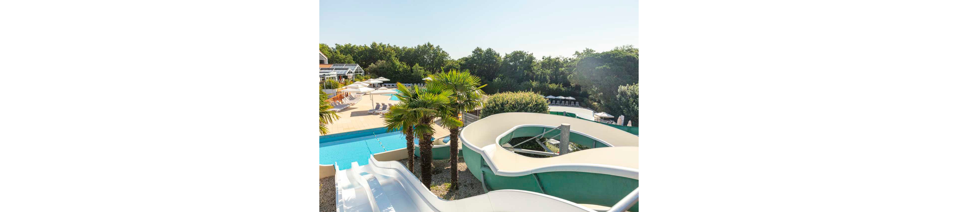 An outdoor water park features large curving water slides leading into a bright blue pool. Palm trees and surrounding greenery create a tropical atmosphere around the aquatic area. Sun loungers and umbrellas line the poolside deck, suggesting a relaxing vacation setting. The clear sky and bright sunlight emphasize a warm, summery day at the resort.