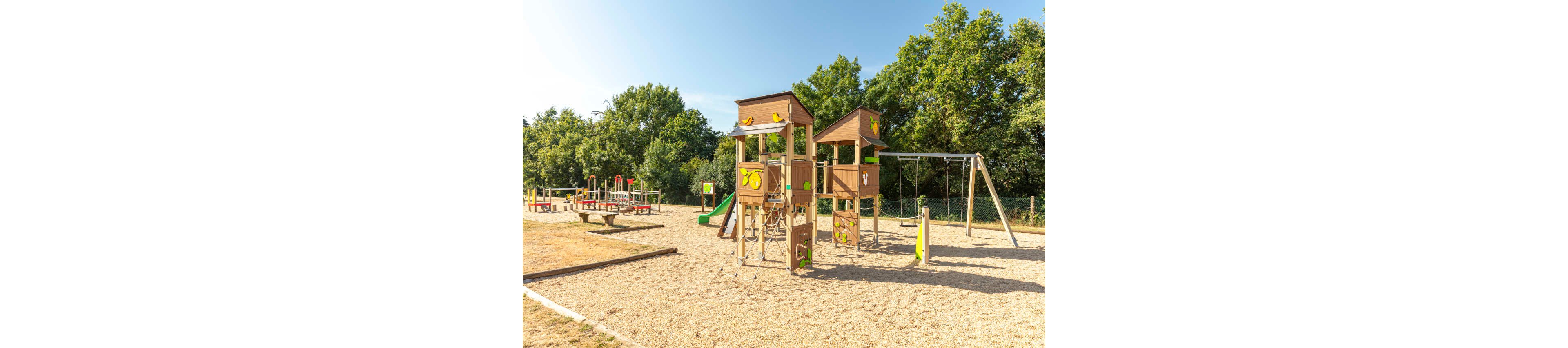 An empty outdoor playground features wooden climbing towers with slides and ladders on a bright sunny day. The play structures stand on a gravel or sand surface surrounded by green trees and grass. Swings, balance equipment, and colorful play elements are visible in the background. The scene suggests a safe recreational area for children in a park or schoolyard.