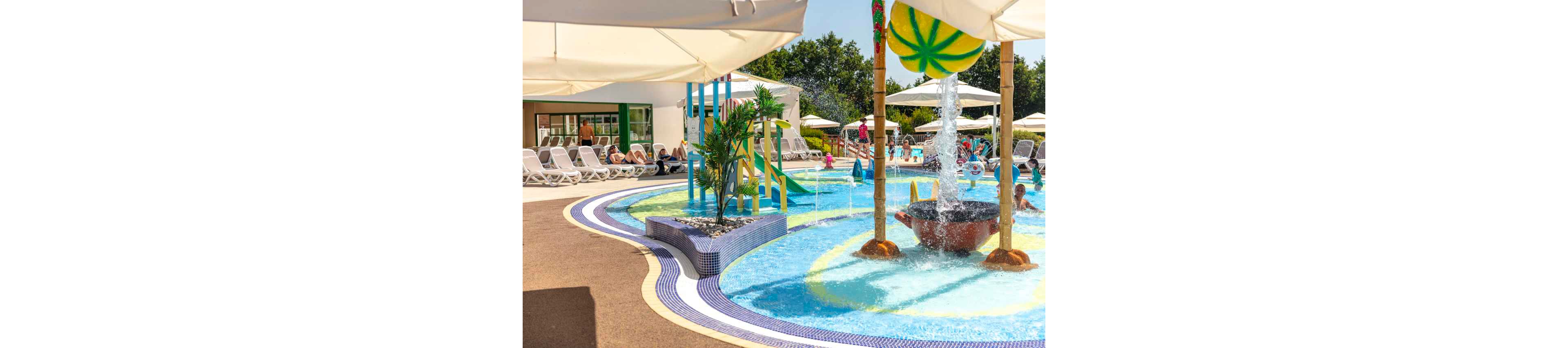 A vibrant children’s splash pool features shallow water, playful slides, and a large overhead water feature. Lounge chairs and umbrellas surround the pool where adults relax in the sun. Bright colors, curved pool edges, and tropical plants create a cheerful vacation atmosphere. The scene suggests a family-friendly resort on a warm, sunny day.