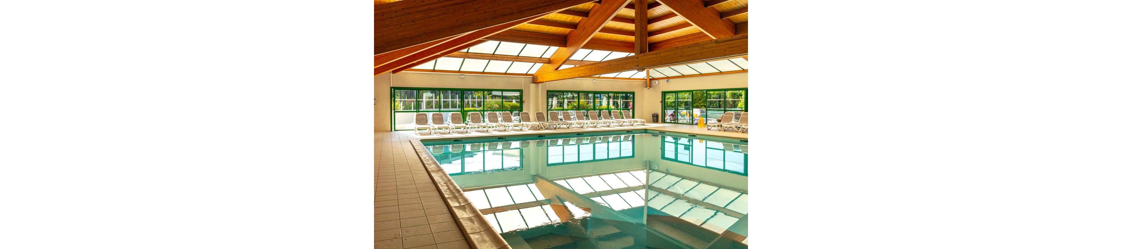 Spacious indoor swimming pool inside a sunlit leisure facility. Numerous white lounge chairs line the far side of the pool, facing large windows that overlook greenery outside. A high wooden beam ceiling and skylights allow natural light to reflect on the calm water surface. The clean tiled floor and empty chairs suggest a peaceful, relaxing atmosphere.