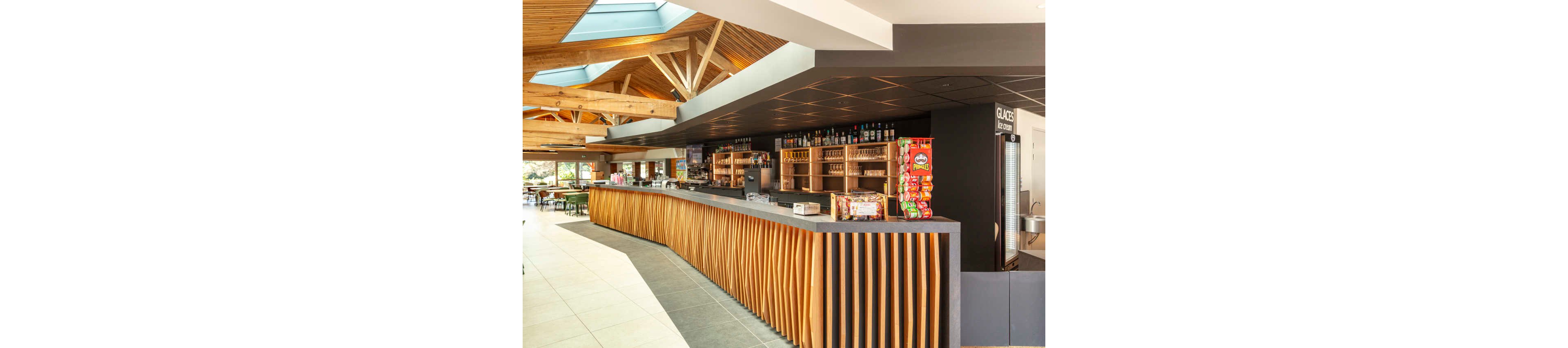 A contemporary cafeteria bar features a long counter with sculpted wooden slats and a dark countertop. Behind the bar, shelves are stocked with bottles, snacks, and refrigerated drinks. Bright natural light enters through large skylights in the wooden ceiling, illuminating the spacious interior. The seating area with tables and chairs is visible in the background, creating an inviting dining atmosphere.