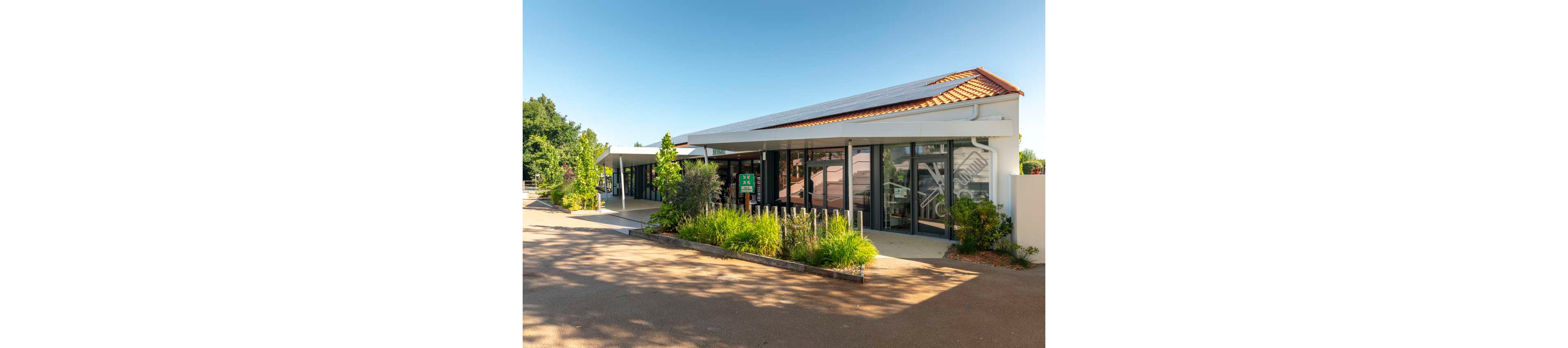A contemporary single-story building with large glass windows is shown on a sunny day. The structure features a sloped tiled roof and a clean white exterior. In front of the building, there is a landscaped garden with green shrubs and plants bordered by a low wooden fence. A wide paved walkway or driveway runs alongside the building, casting long shadows in the bright light.