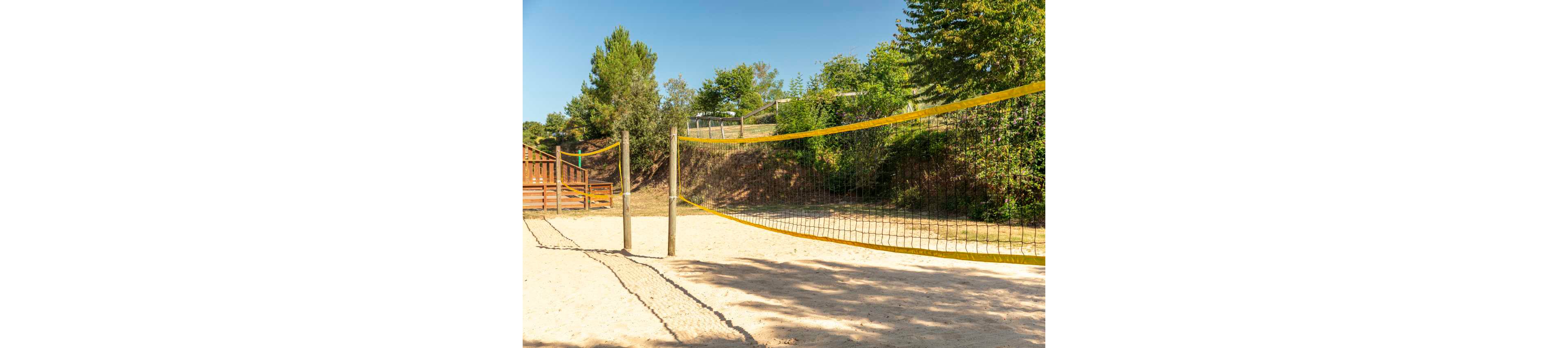 An empty sand volleyball court is set up outdoors with a yellow net stretched between wooden posts. The court is surrounded by green trees and grassy slopes under a clear, sunny sky. Wooden structures and fencing are visible in the background, suggesting a recreational park or campground setting. The bright daylight and clean sand create a warm, inviting atmosphere for outdoor sports.