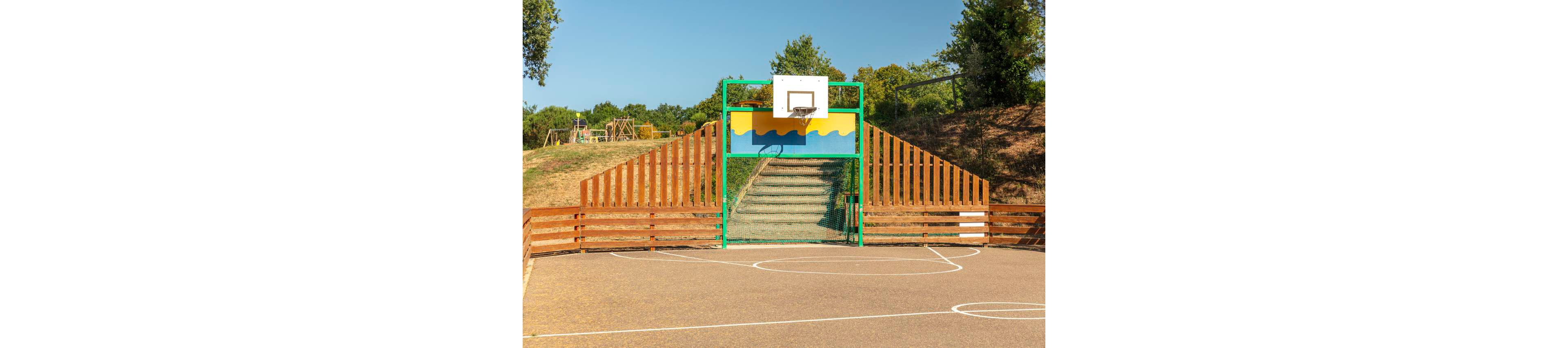 An outdoor basketball court is shown with a single hoop mounted above a colorful painted backboard. Behind the hoop, a fenced staircase leads up between slanted wooden railings on a grassy slope. The court surface is marked with white lines for play, and the area is surrounded by trees and greenery. In the background, a wooden playground structure is visible on the hill.