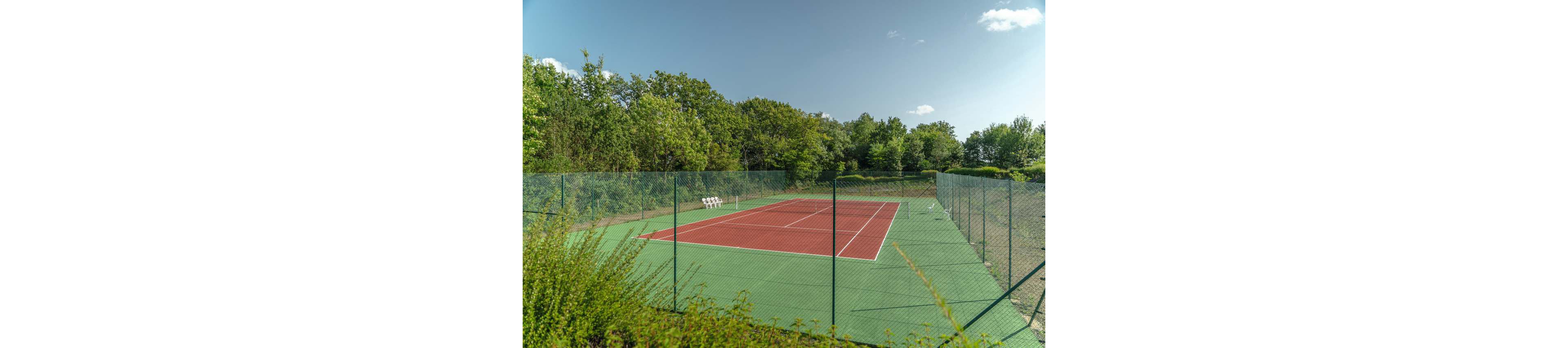 An empty red and green tennis court is surrounded by a tall chain-link fence. The court sits in a lush, green landscape with dense trees and bushes on all sides. A small stand with tennis balls is visible near the far baseline. The scene is lit by bright daylight under a mostly clear blue sky with a few scattered clouds, creating a calm and peaceful atmosphere.