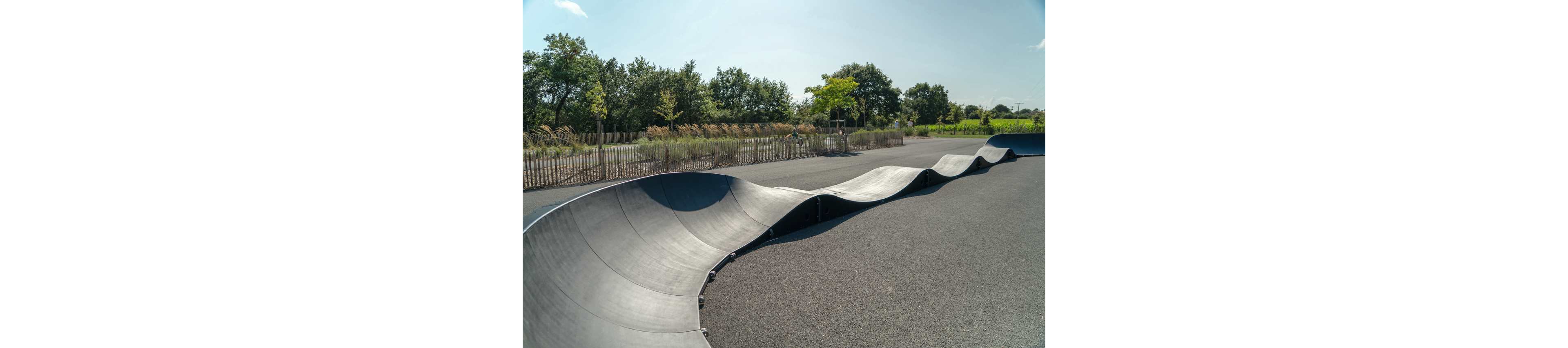 A smooth black pump track with flowing curves winds across an open paved area. The track is set in an outdoor park bordered by wooden fencing and ornamental grasses. Green trees and a bright blue sky with light clouds create a calm, sunny atmosphere. The scene is empty of people, emphasizing the sculptural shape of the track and the spacious surroundings.
