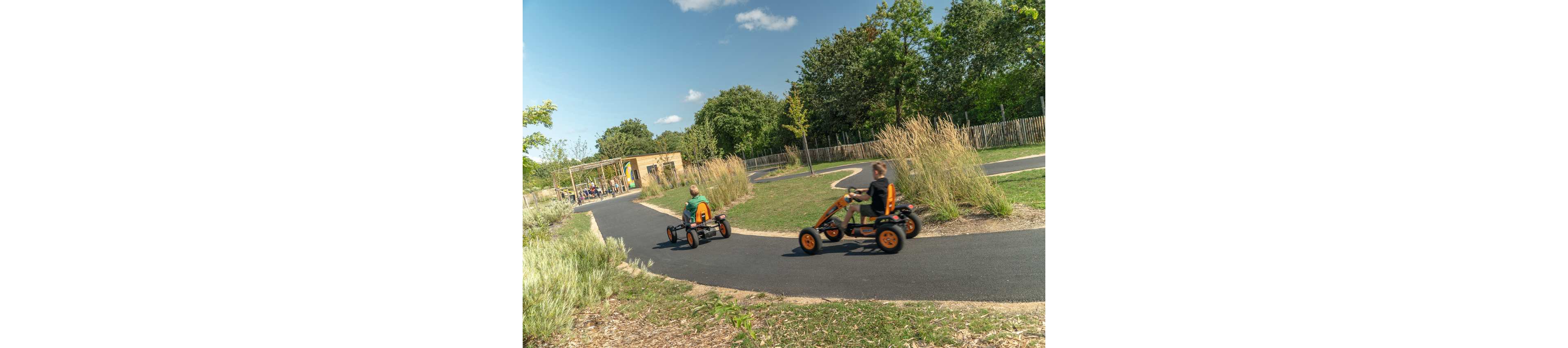 Two children ride orange pedal go-karts along a winding paved track in an outdoor park. Tall grasses and landscaped greenery border the path, creating a natural setting. A wooden building and additional people are visible in the background near the track entrance. The scene is bright and sunny, suggesting an active and playful atmosphere.