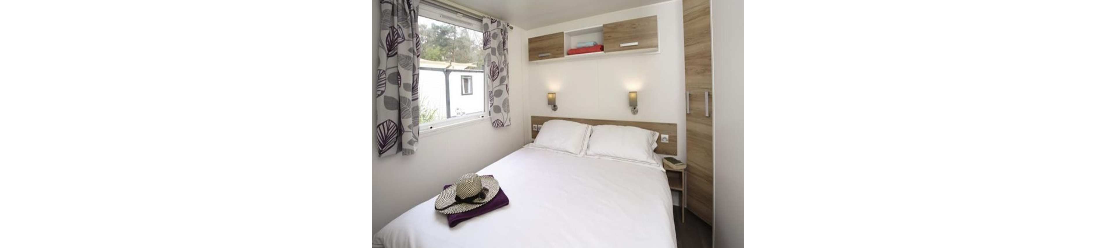 A small, modern bedroom features a neatly made double bed with white linens. A straw hat and folded clothing rest at the foot of the bed, suggesting a vacation or weekend stay. Built-in wooden cabinets and wall-mounted reading lights frame the bed, adding functional storage and warm lighting. A window with patterned curtains lets in natural light and offers a view of trees and neighboring structures.