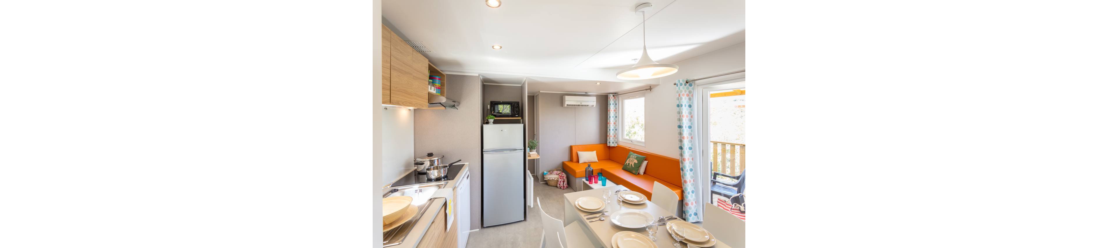 Compact open-plan kitchen and living area inside a modern mobile home. A small dining table is set with plates and glasses beside a bright orange sofa and colorful cushions. The kitchenette includes a stove with pots, a sink, and a tall fridge with a microwave on top. Large windows with patterned curtains and a glass door let in natural light, creating a cheerful vacation atmosphere.