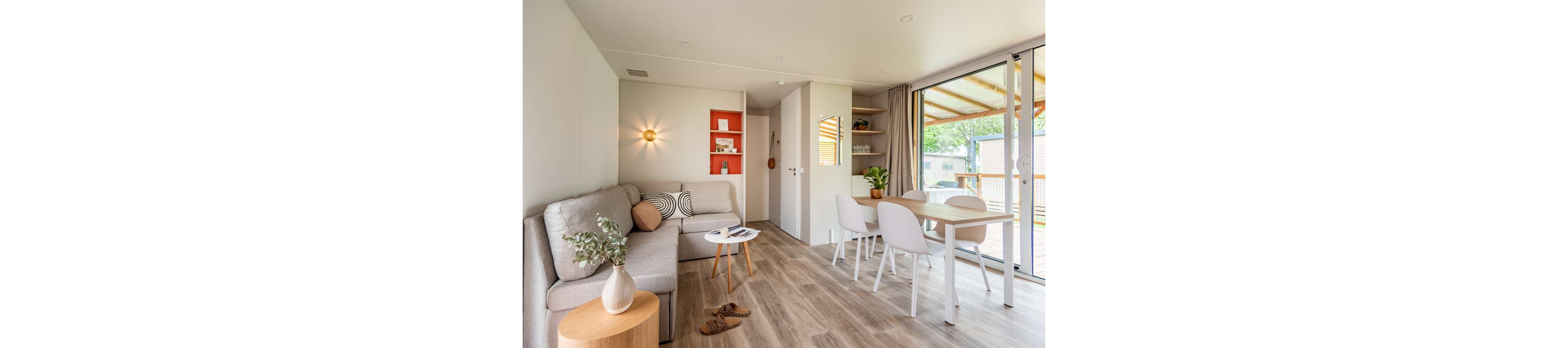 Contemporary open-plan living space features a light gray sofa and small round side table with a vase of greenery. A dining table with white chairs sits beside large sliding glass doors leading to a covered terrace. Built-in shelving with orange accents and minimal decor adds a touch of color to the neutral room. Wooden flooring, soft lighting, and simple furnishings create a cozy, inviting atmosphere.