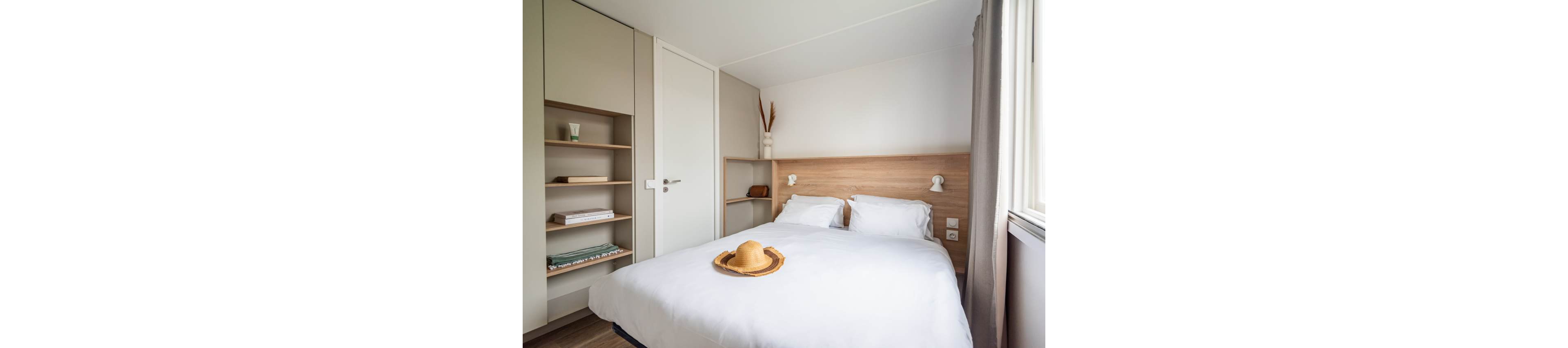 A compact modern bedroom features a neatly made double bed with white linens and a straw hat placed at the foot. Light wood tones on the headboard and floor create a warm, minimalist atmosphere. Built-in shelves and a wardrobe provide storage space, holding a few books and personal items. Natural light streams in through a window with curtains, giving the room an airy and inviting feel.