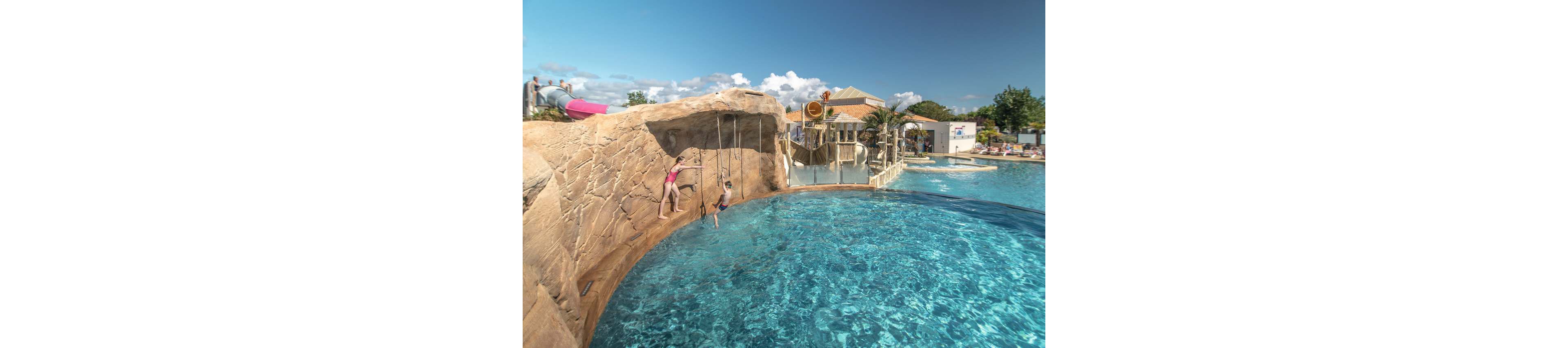Two children play on hanging ropes above a large outdoor swimming pool. The pool is designed with artificial rock formations and clear blue water. In the background, there is a water slide, wooden play structures, and resort buildings under a bright sky with scattered clouds. The scene suggests a family-friendly vacation destination focused on water activities and fun.