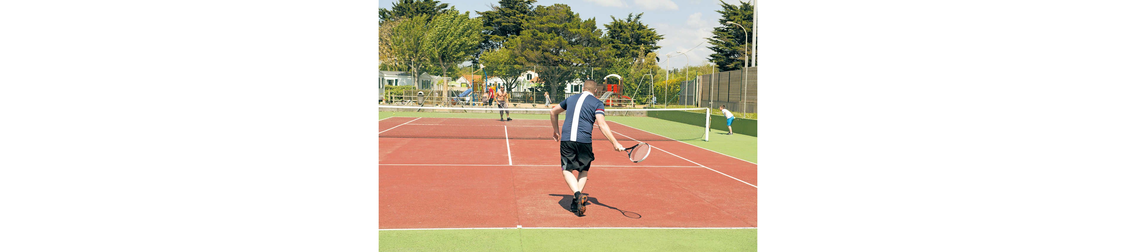A man is playing tennis on an outdoor hard court, captured from behind as he moves toward the net. Other players are visible on the opposite side of the court and on a neighboring court. The scene is set in a recreational area with trees, buildings, and a playground in the background. Bright daylight and clear skies create a casual, active atmosphere.