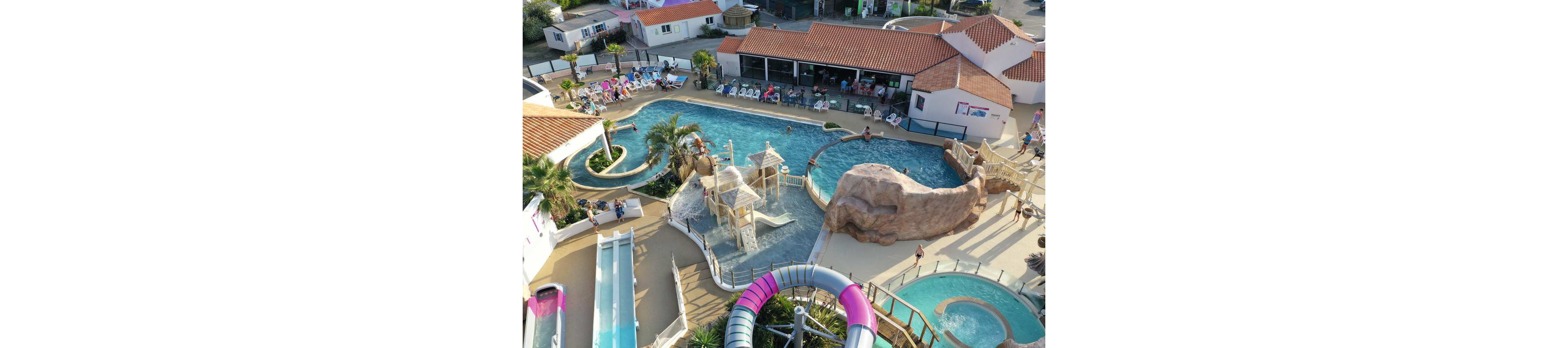 A large outdoor water park is captured from above, showing multiple pools and winding water slides. Families and children play in shallow splash areas with small towers, bridges, and interactive water features. A rock-like structure and palm trees add a tropical theme to the scene. Sunbathers relax on lounge chairs around the pools, while a building with a terrace overlooks the aquatic area.