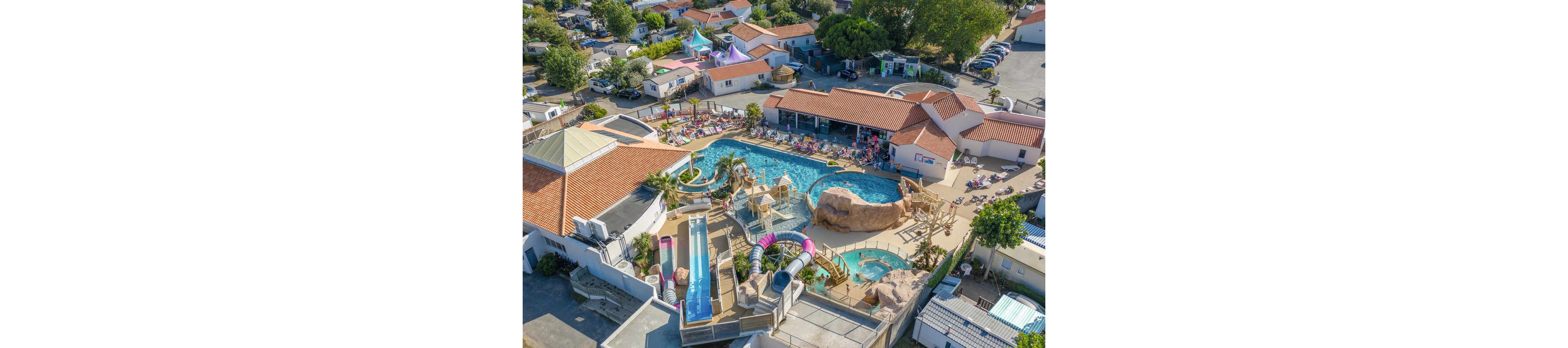A large outdoor swimming pool complex is seen from above at a busy holiday resort. Families and children relax on sun loungers and enjoy multiple pools, slides, and water play structures. The scene includes rock features, palm trees, and colorful tube slides creating a fun, tropical atmosphere. Surrounding the waterpark are low-rise buildings with red-tiled roofs and nearby campsites, emphasizing a vacation destination.