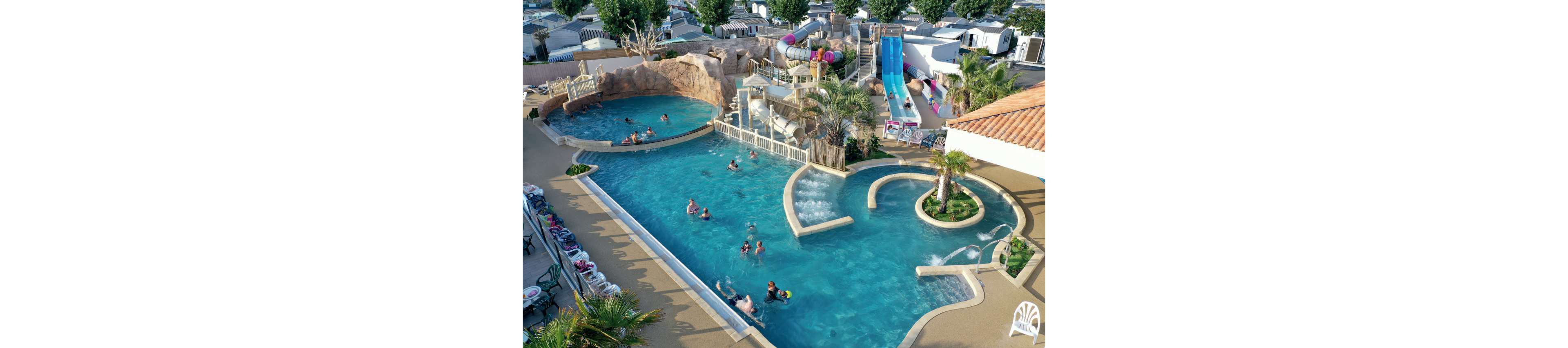 A large outdoor swimming pool complex is shown from above at a family resort. Multiple pools, curved channels, and bubbling water features create a playful aquatic environment. Colorful water slides and a small play structure rise above the pools, with people swimming and relaxing in the water. Surrounding the pools are lounge chairs, palm trees, and nearby cabins or mobile homes, suggesting a holiday campground setting.