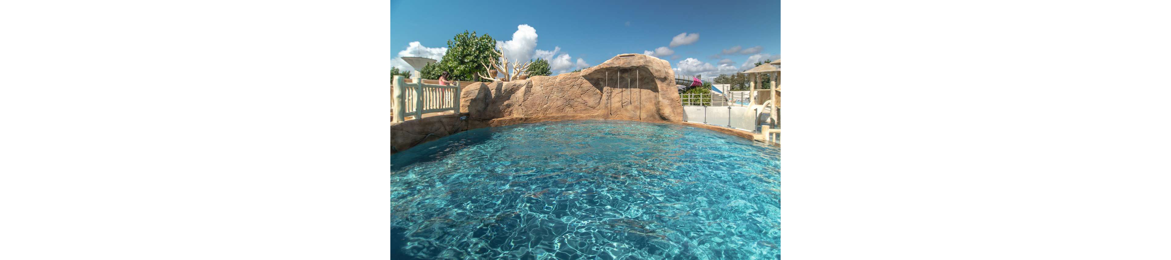 Children play in a large outdoor swimming pool at a resort-style water park. One child swims in the clear blue water while others stand and climb on a rocky structure with metal ladders. The pool is surrounded by railings, trees, and lounge areas under a bright blue sky with scattered clouds. The scene feels relaxed and fun, capturing a sunny vacation atmosphere.
