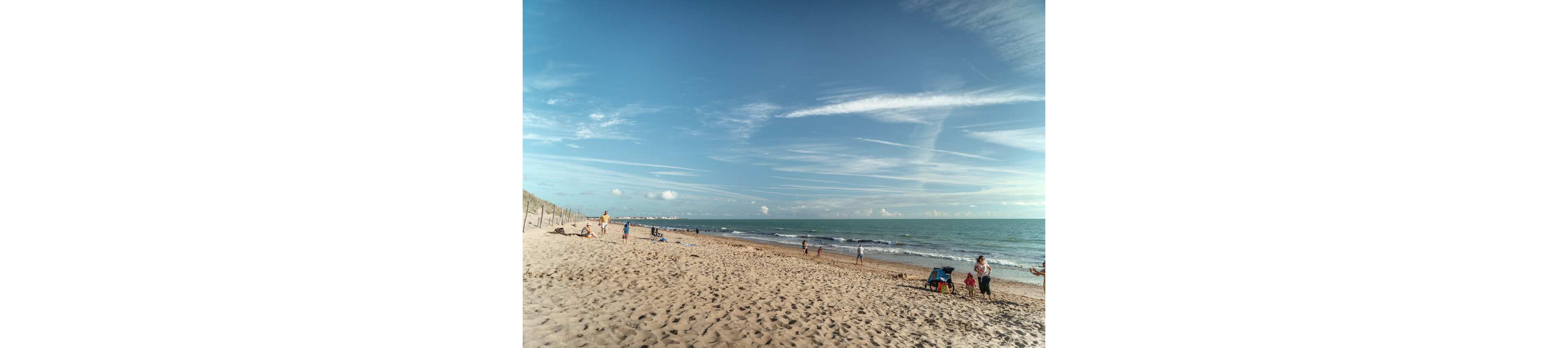Several people and families relax along a wide sandy beach beside a calm sea. Some individuals walk near the shoreline while others sit on beach chairs or towels closer to the dunes. The sky is mostly clear with scattered clouds and long streaks of high-altitude clouds stretching across the horizon. The overall scene feels peaceful and leisurely on a bright day by the ocean.