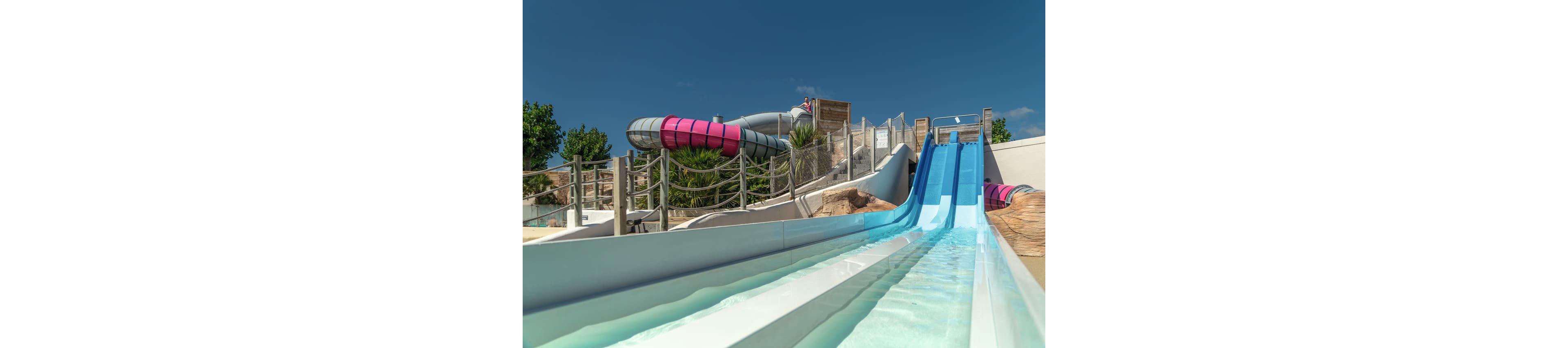 A set of bright blue and white water slides stretches down toward the viewer in an outdoor water park. In the background, a large enclosed tube slide with pink and gray sections curves above the open flumes. The scene is sunny with a clear blue sky, palm trees, and wooden fencing surrounding the attraction. No people are present, emphasizing the structure and clean, inviting water.
