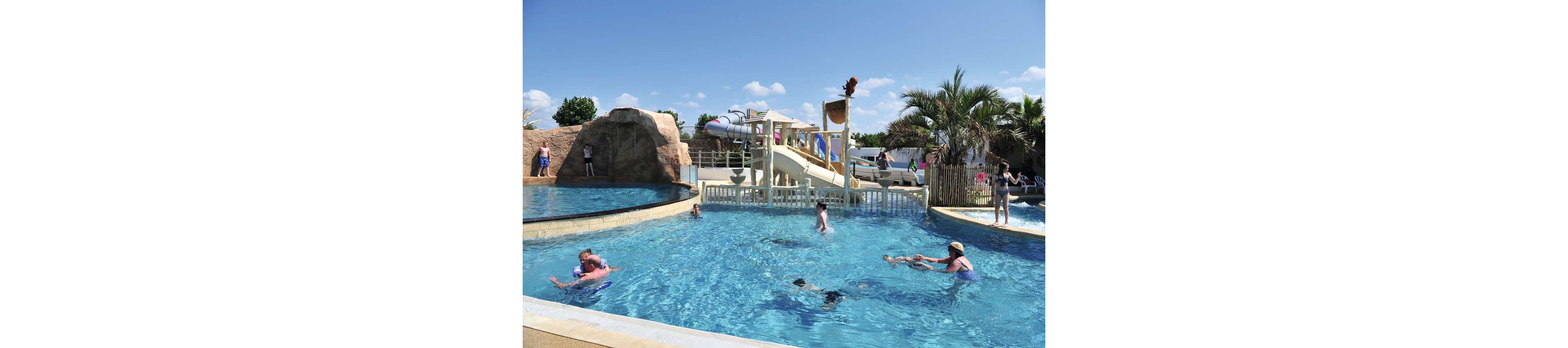 An outdoor swimming pool at a water park is filled with families enjoying a sunny day. Children and adults play in the clear blue water while others relax near the pool edge. A large rock feature, water slides, and a splash bucket structure create a fun, resort-like atmosphere. Palm trees, lounge chairs, and bright skies emphasize a warm, vacation mood.