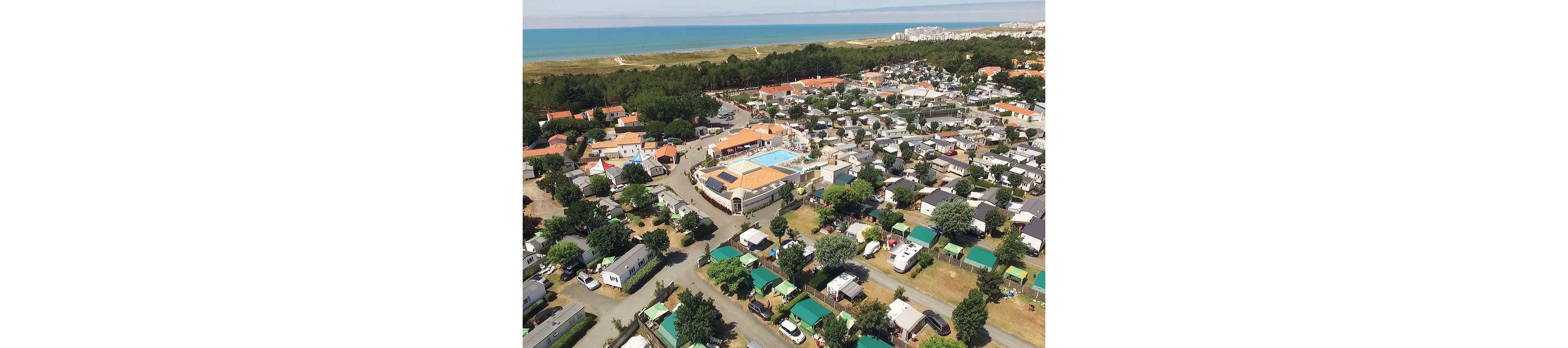 A large holiday campsite with numerous caravans and mobile homes is seen from above near a coastline. At the center of the site, a swimming pool complex and main building stand out among the rows of pitches. Green trees and hedges separate the plots, creating a structured yet natural layout. In the distance, sandy dunes and the sea stretch along the horizon, with a town visible further along the coast.