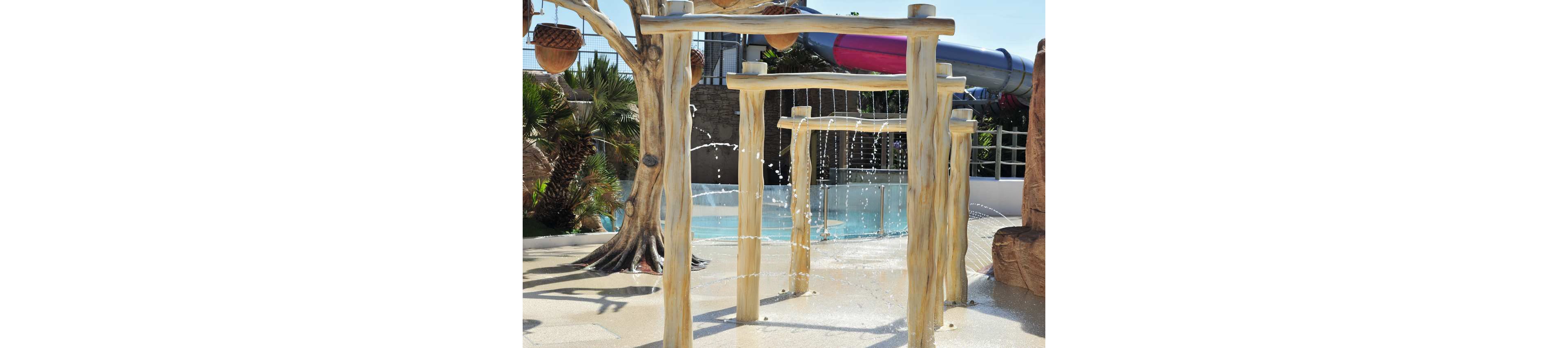 Children in swimsuits play under cascading water jets at an outdoor splash park. Wooden-style arches frame the water features as kids run through the streams. A pool and large water slide are visible in the background, suggesting a family-friendly water park. Bright sunlight and clear skies create a warm, summery atmosphere.