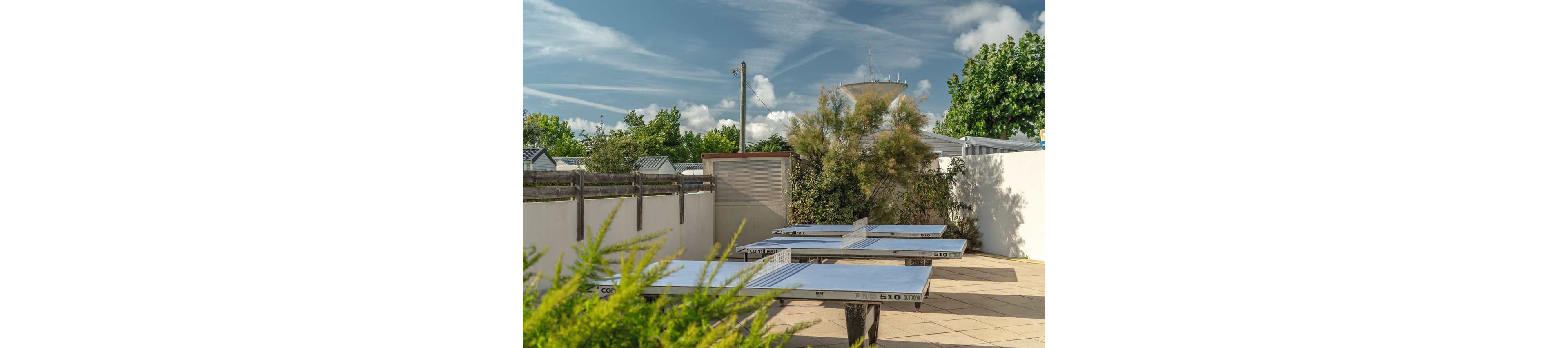 A group of adults and children play table tennis on outdoor tables in a sunny recreational area. The scene appears to be in a holiday park or campground courtyard, surrounded by fences, trees, and small buildings. Bright daylight and blue skies with scattered clouds create a cheerful summer atmosphere. The players are casually dressed, suggesting a relaxed family vacation activity.