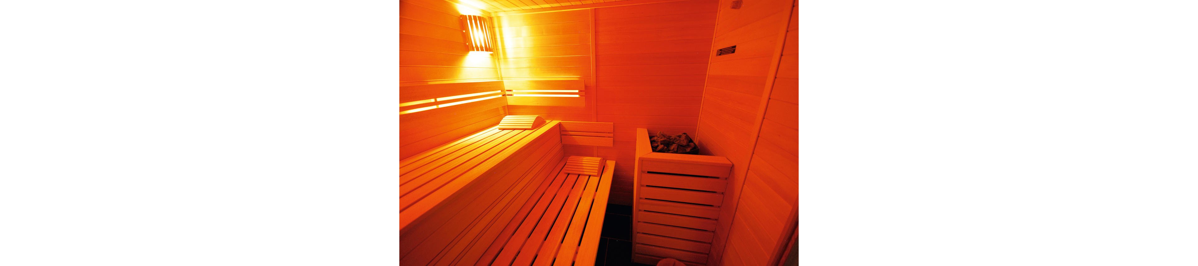 Image of a cozy wooden sauna interior with tiered benches. A heater filled with sauna stones is positioned in the corner, enclosed by wooden slats. The space is illuminated by warm orange lighting, creating a relaxing atmosphere. Smooth wooden paneling covers the walls and ceiling, emphasizing a clean and minimalist spa environment.