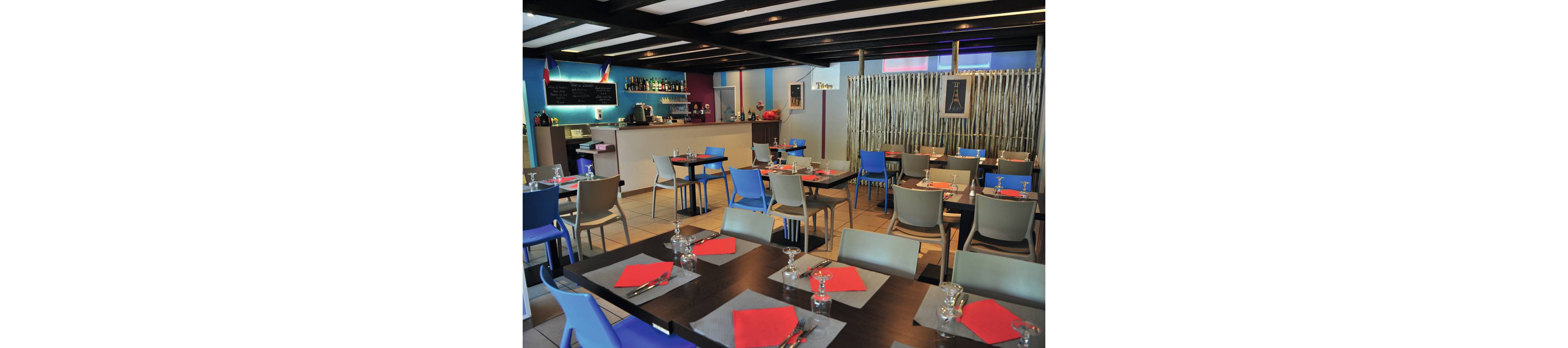 A contemporary restaurant dining area features neatly arranged tables with red napkins and glassware. Colorful chairs in blue and gray surround dark wooden tables, creating a vibrant yet casual atmosphere. In the background, a small bar area with bottles and a chalkboard menu adds to the cozy setting. Soft lighting and decorative wall elements give the space a relaxed, welcoming feel.