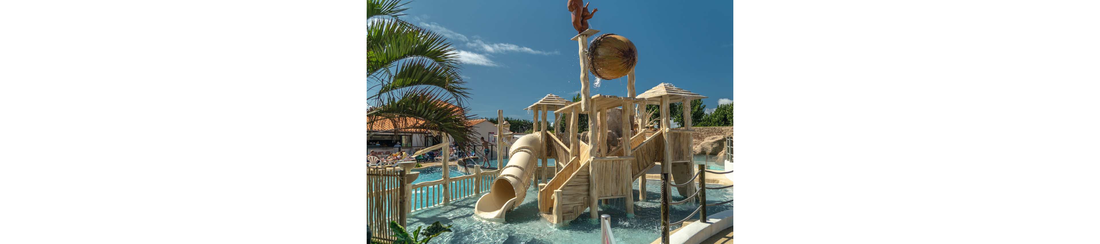 A tropical-style wooden water playground stands in a shallow pool under a bright blue sky. The structure features slides, stairs, and platforms surrounded by clear water and safety railings. A large bucket at the top pours water down as part of the splash attraction. Palm trees and sunbathers are visible in the background, creating a relaxed vacation atmosphere.